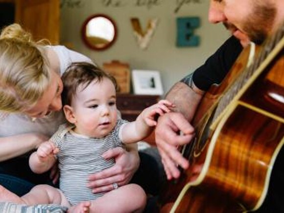 Mom holding baby listening to dad playing guitar
